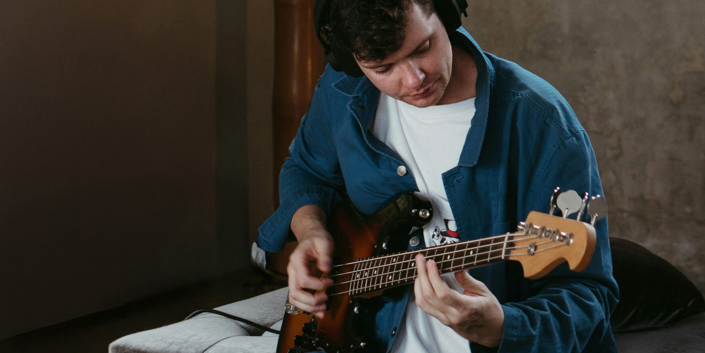 A musician in headphones playing a bass guitar on a studio sofa.