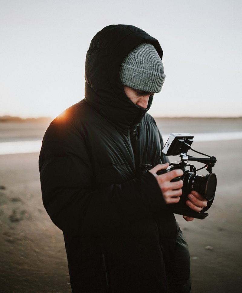 A filmmaker in a hooded coat checking their camera on a beach at sunrise.