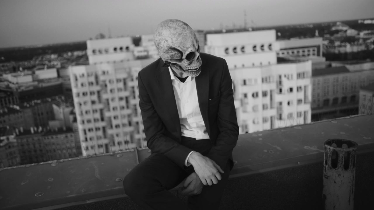 Figure in Skull Mask Sitting on City Rooftop