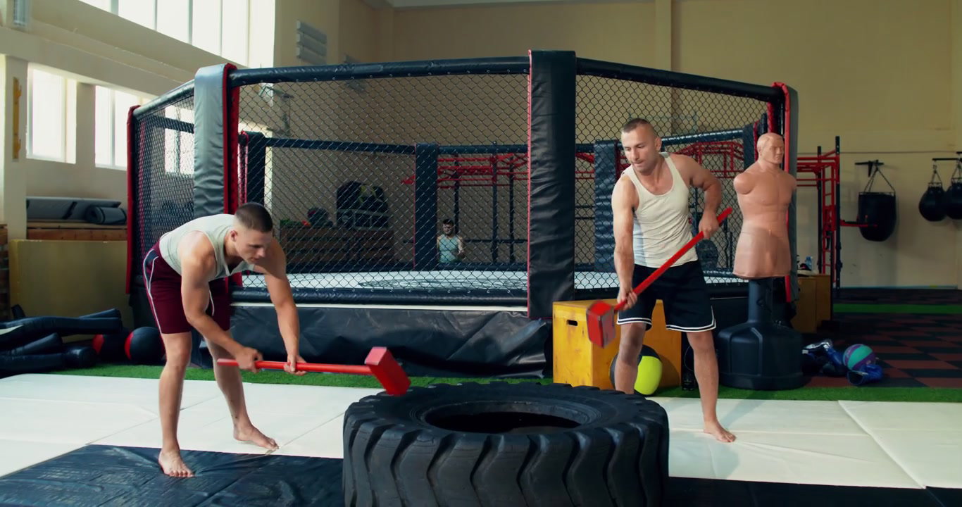 Two Men Hitting Tire with Sledgehammers in Gym